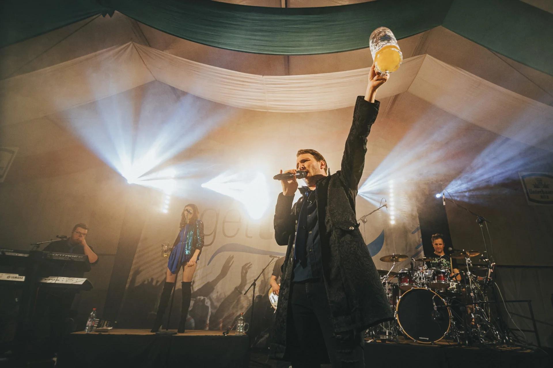 Ein Musiker auf einer Bühne hält ein Mikrofon in der einen Hand und ein Glas mit Getränk in der anderen. Im Hintergrund spielen weitere Musiker, darunter eine weibliche Sängerin und ein Schlagzeuger. Die Bühne ist beleuchtet mit hellen Lichtstrahlen 