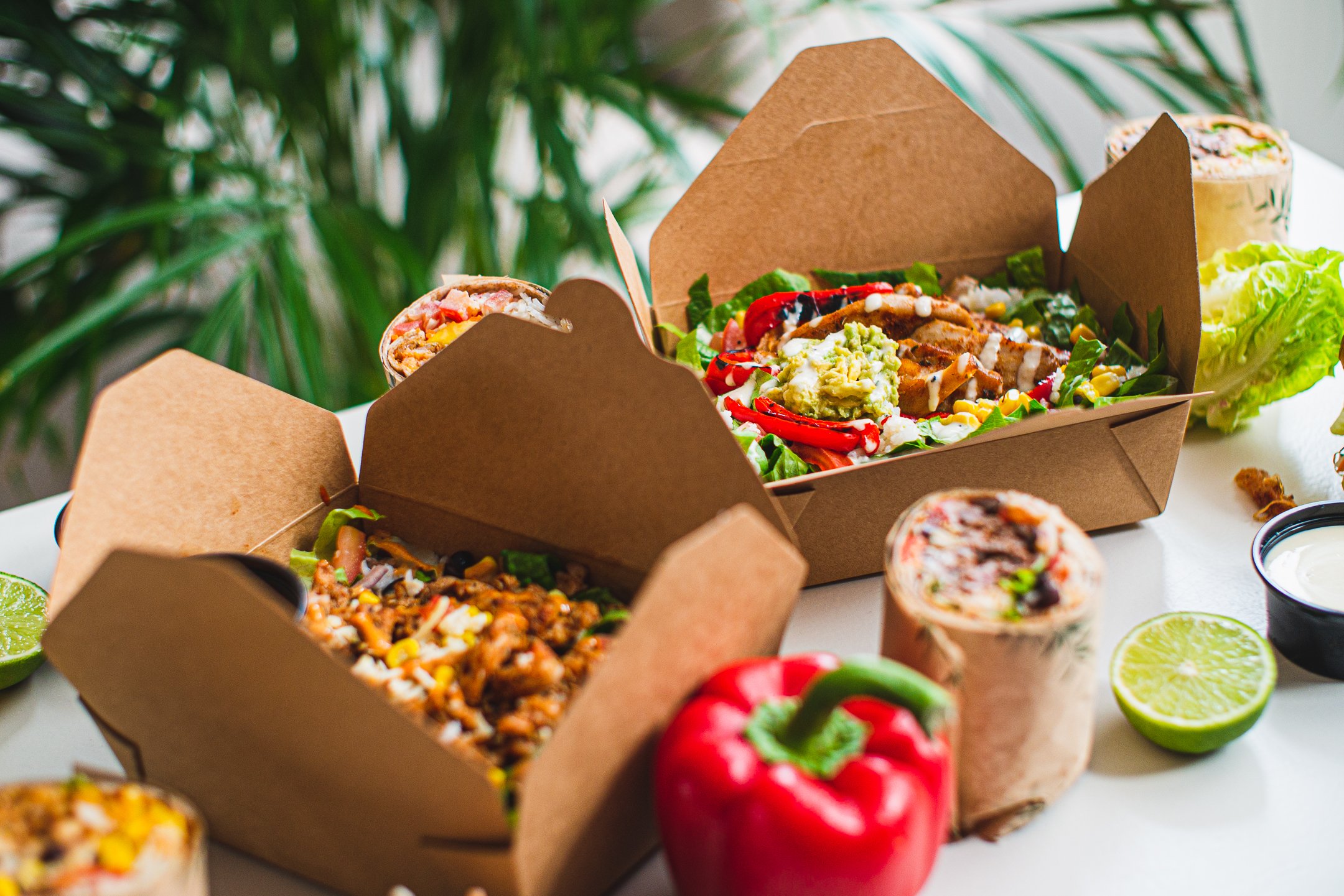 Mexicaanse maaltijd in kartonnen bakjes met salades, bowls, wraps, limoen en diverse groenten zoals paprika en sla.