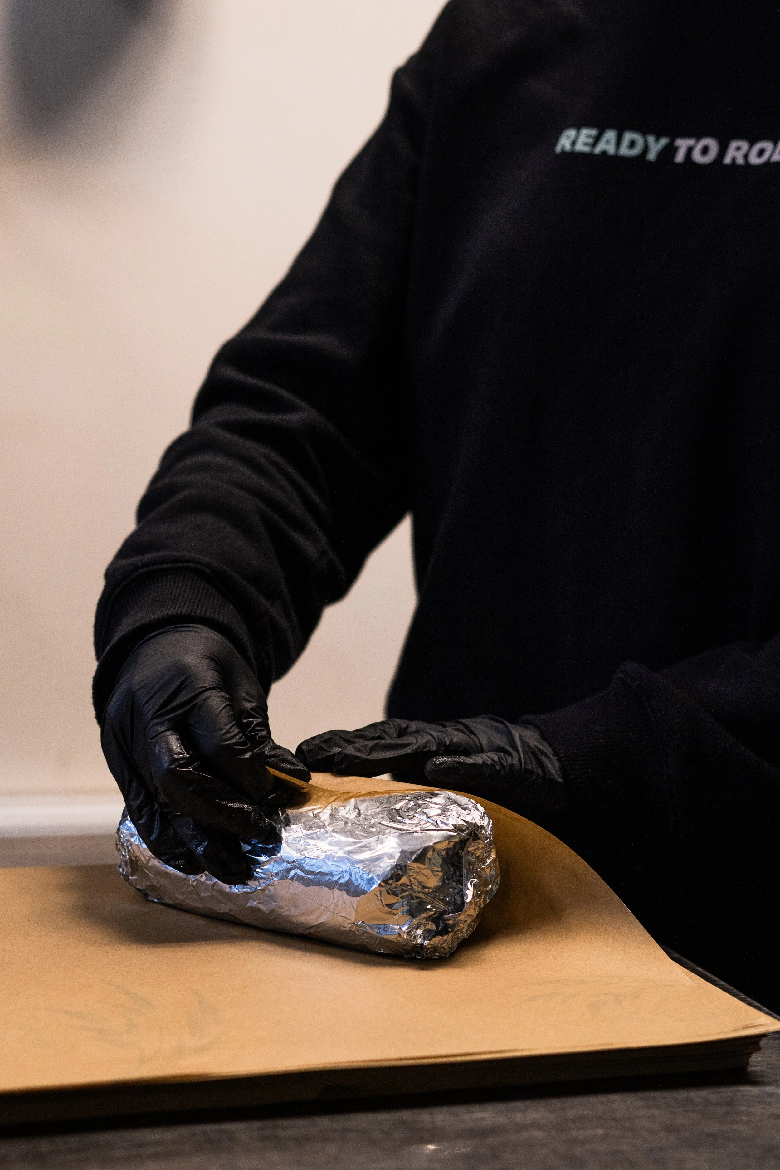Persoon met zwarte handschoenen wikkelt een burrito in aluminiumfolie op een tafel.