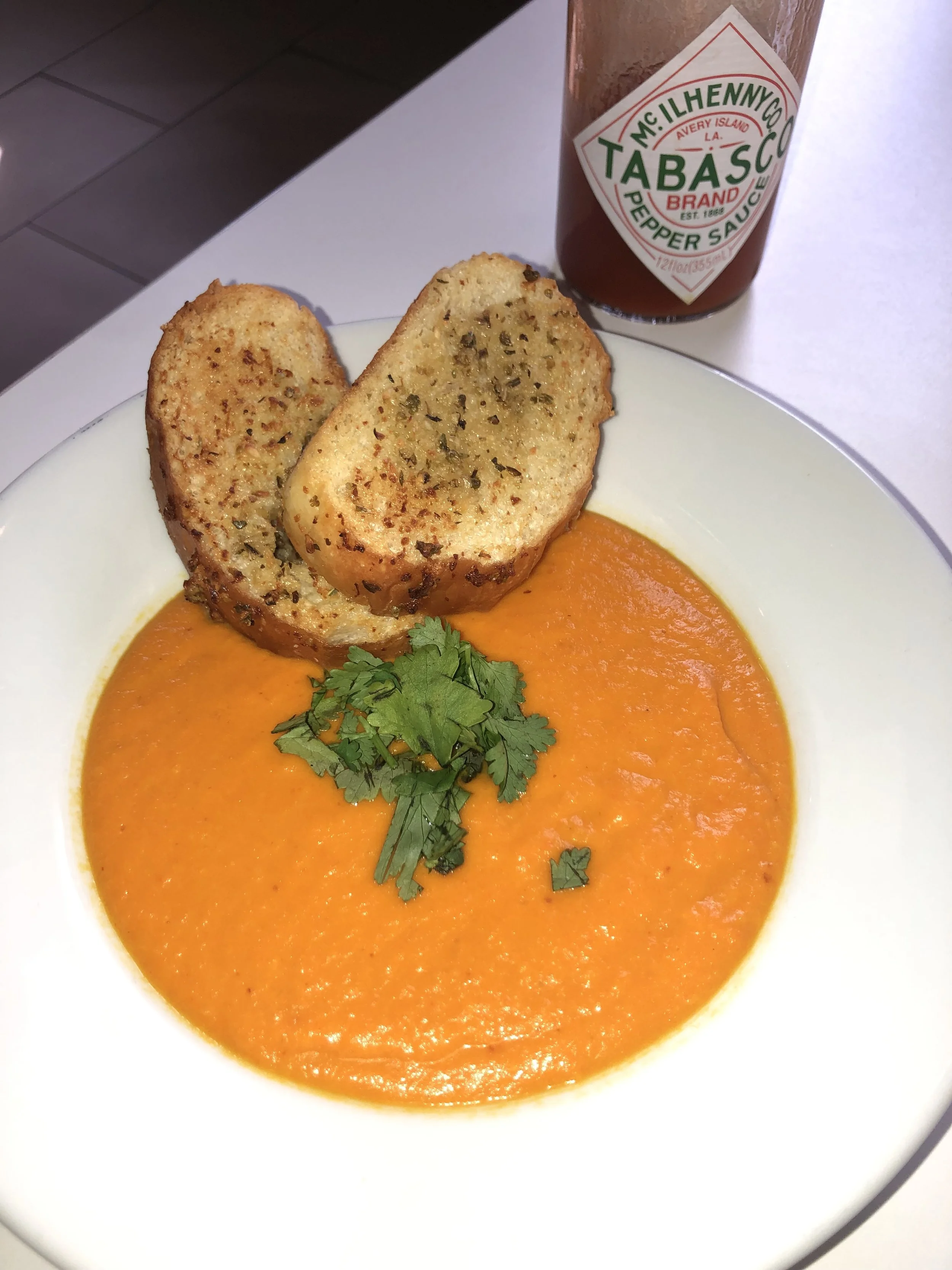 A bowl of bright orange tomato soup topped with cilantro, served with two slices of garlic bread, and a bottle of Tabasco pepper sauce in the background.