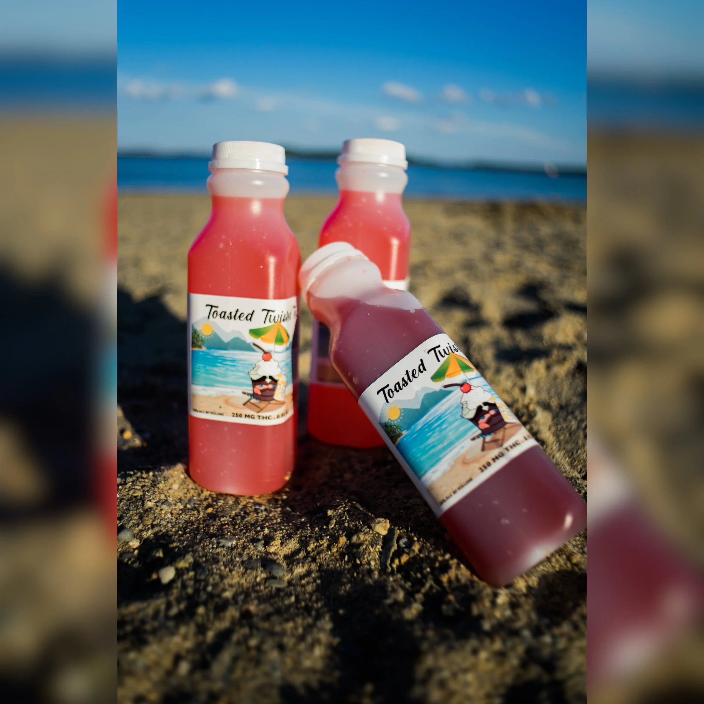 Three bottles of pink liquid sunscreen or lotion labeled "Toasted Twists" placed on the sandy beach with the ocean and blue sky in the background.
