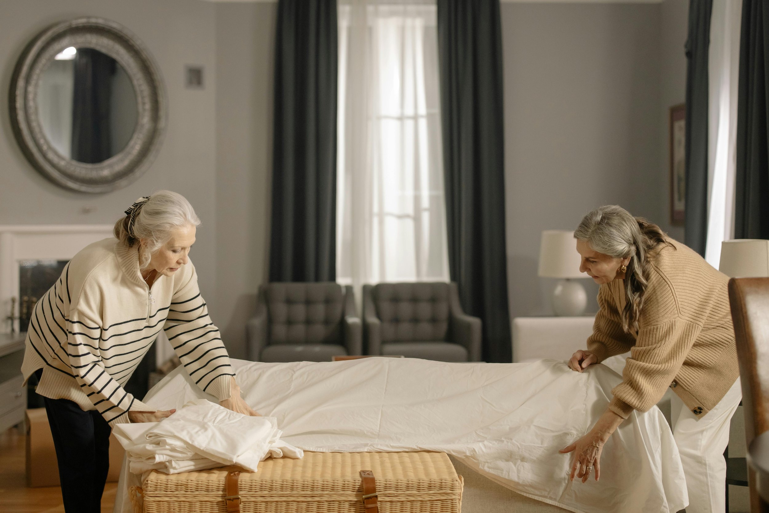 Two elderly women are making a bed together in a well-lit bedroom with a fireplace and a large mirror on the wall.