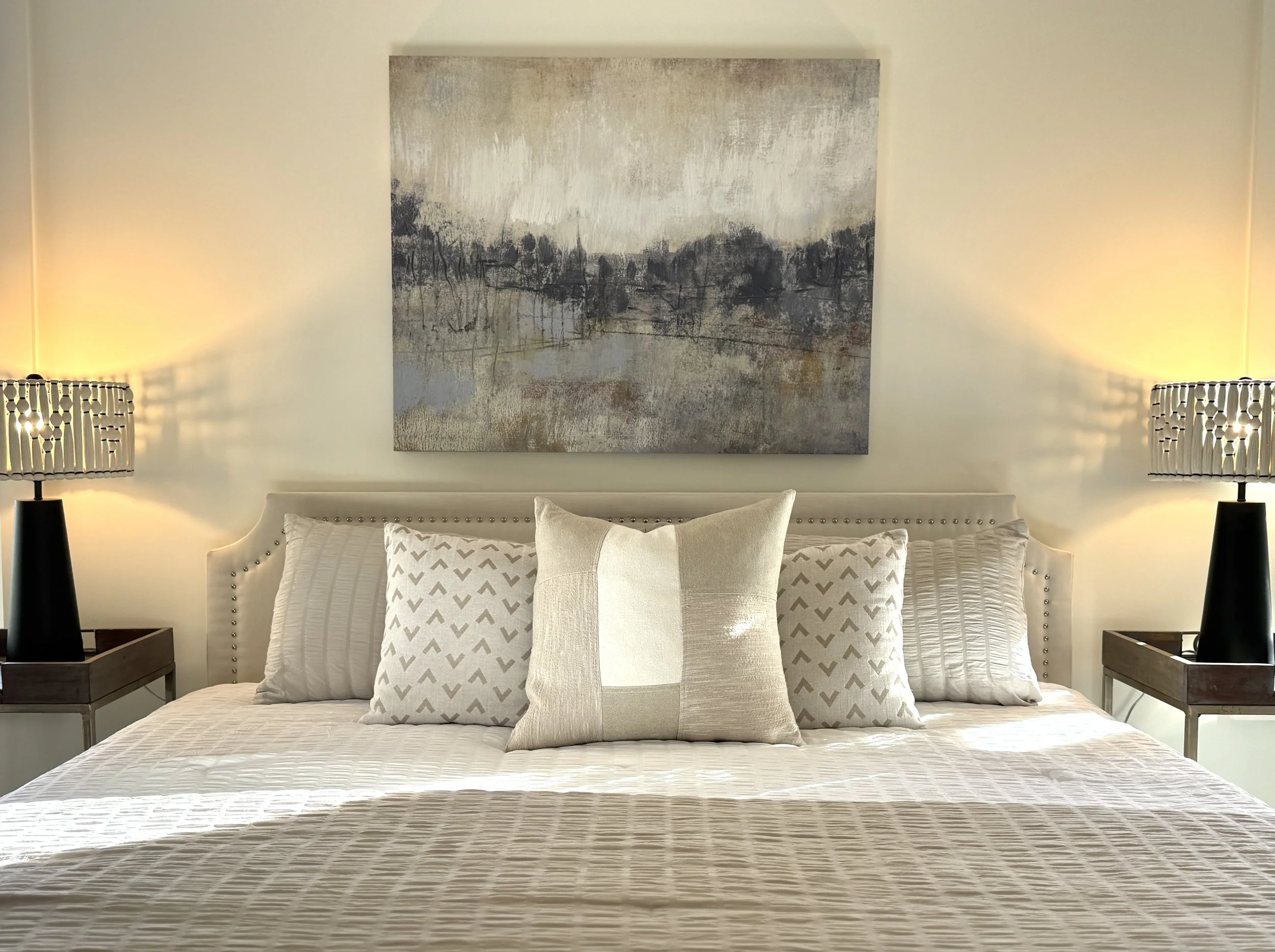 A neatly made bed with white bedding, four decorative pillows, and a headboard with nailhead trim. Above the bed hangs a large abstract landscape painting. There are matching lamps on nightstands on each side of the bed, casting warm light.