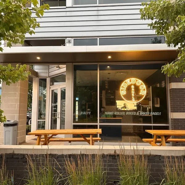 exterior view of a bagel shop with large glass windows, outdoor picnic tables, and a neon sign for Side Door Bagels.