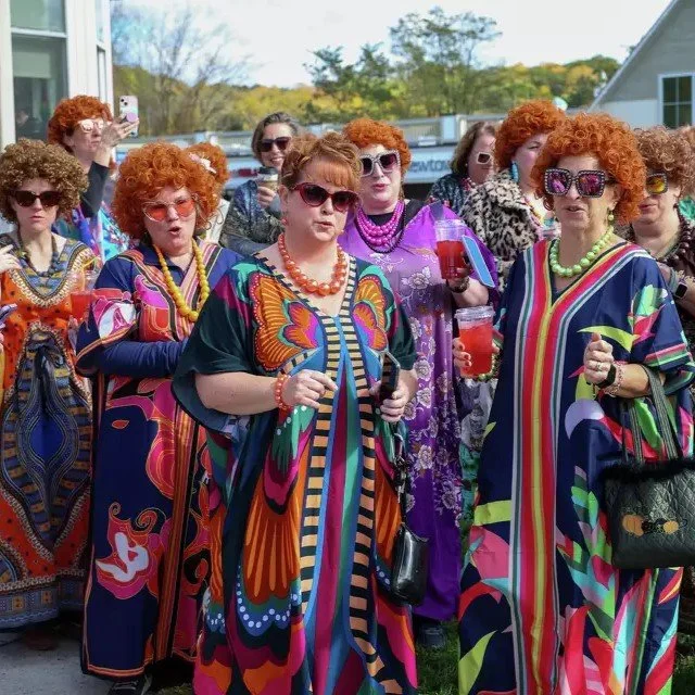 Group of women dressed in colorful, psychedelic-patterned dresses with sunglasses, holding drinks, and outdoor gathering. Mrs. Roper from Three's Company inspired.