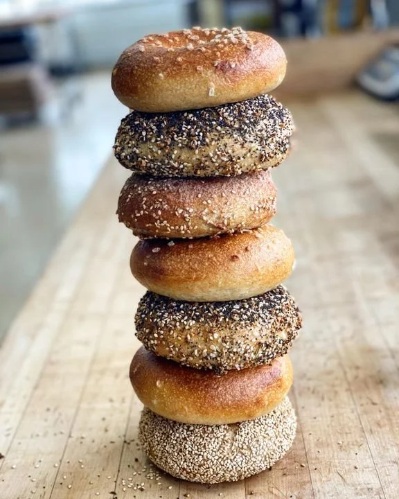 A tall stack of eight assorted bagels on a wooden surface in a cafe or bakery setting.