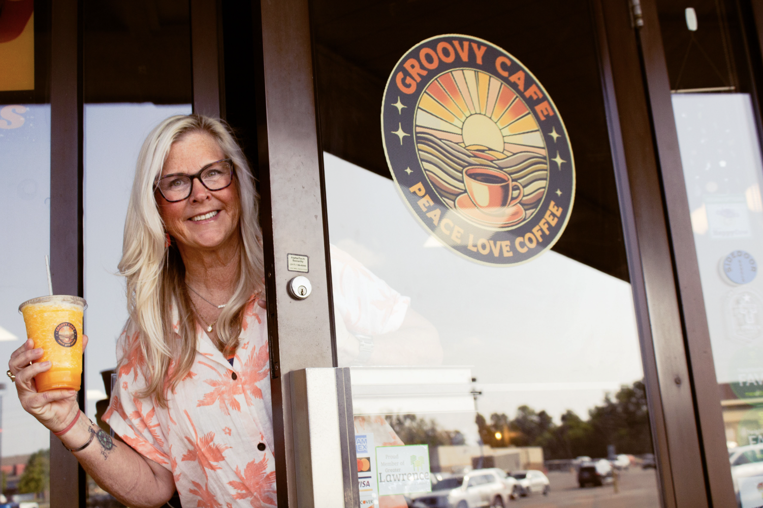 A woman with blonde hair, wearing glasses and a light pink floral shirt, smiling and holding a cold orange-colored drink in a clear plastic cup with a logo, standing in front of a glass door with a sign that says "GROOVY CAFE PEACE LOVE COFFEE."