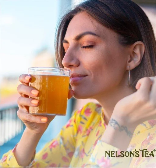 Woman with closed eyes holding a glass of iced tea near her face, wearing a yellow floral shirt, with the Nelson's Tea logo in the corner.