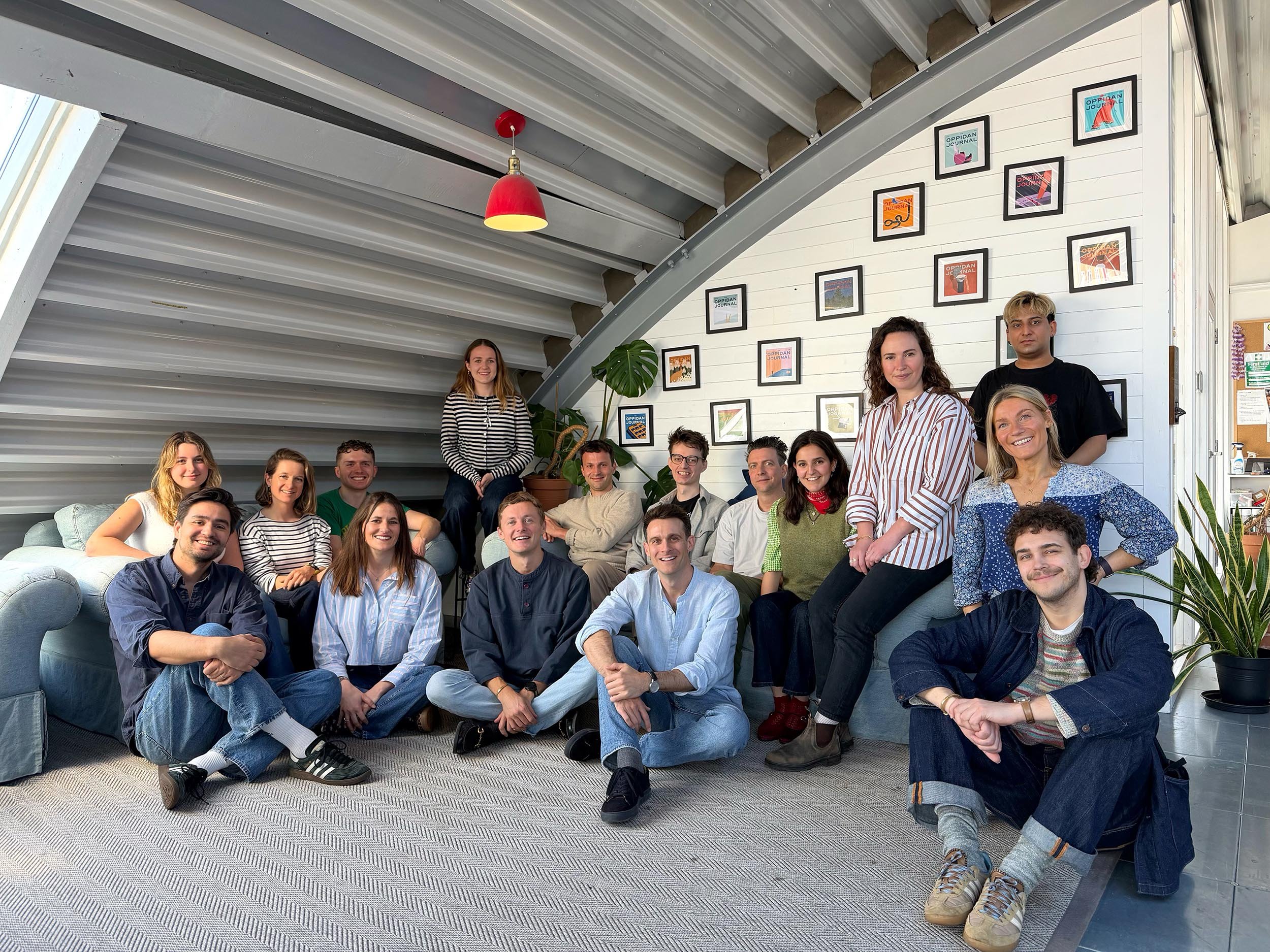 Group of young adults posing together inside a modern room with white walls and artwork, some sitting on a sofa and others standing or sitting on the floor, smiling at the camera.