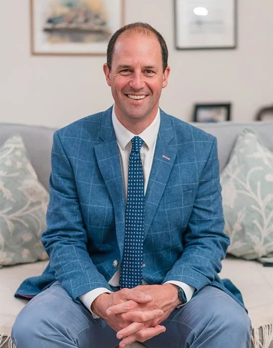A middle-aged man in a suit with a blue tie, smiling, standing inside a warmly lit library with bookshelves in the background.
