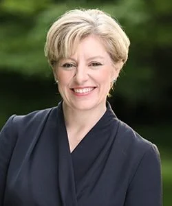 Smiling woman with blonde hair wearing a black blazer and white top standing outdoors near a brick building with large windows.