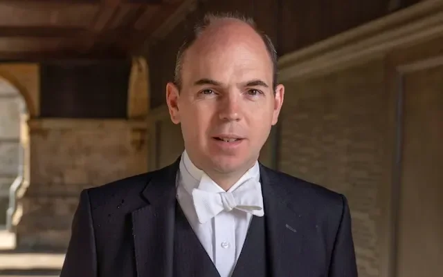 A middle-aged man in a suit with a blue tie, smiling, standing inside a warmly lit library with bookshelves in the background.