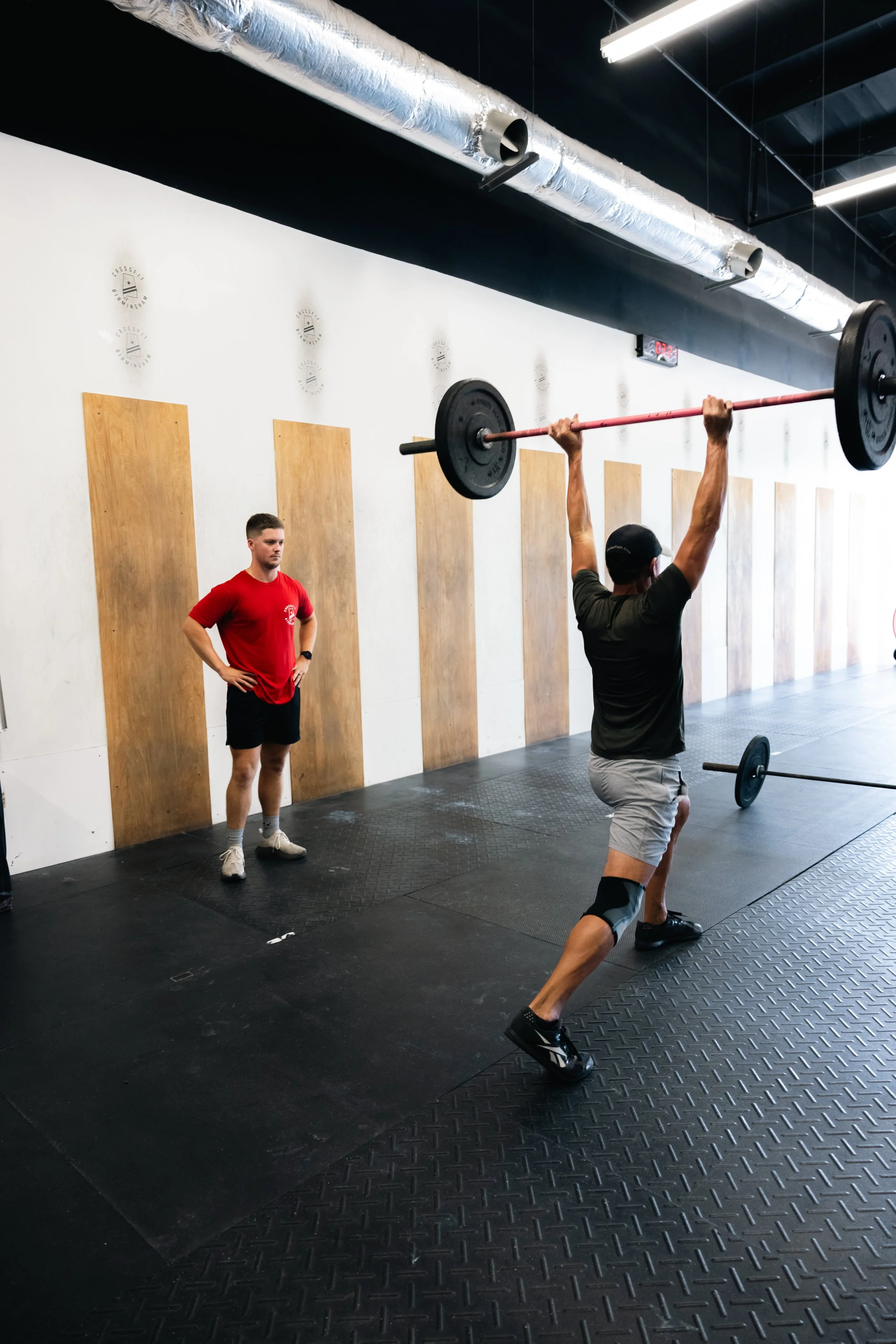A man performing an overhead barbell lift in a gym while a trainer watches.