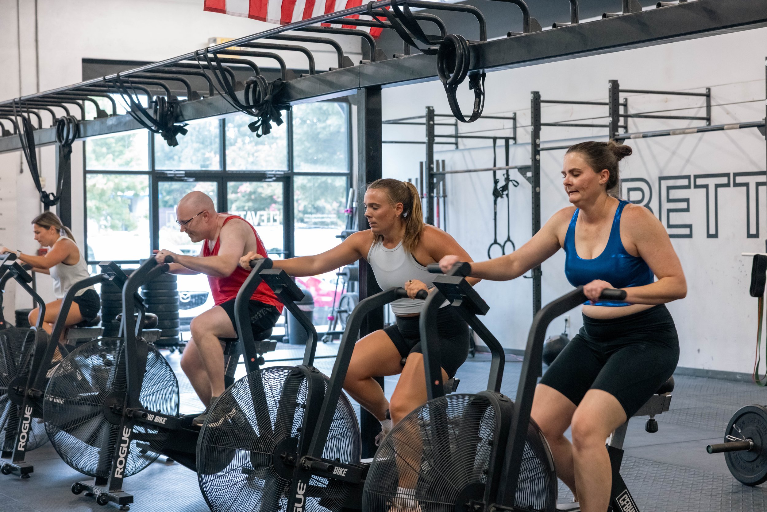 People workout on air bikes in a gym with large windows and fitness equipment in the background.
