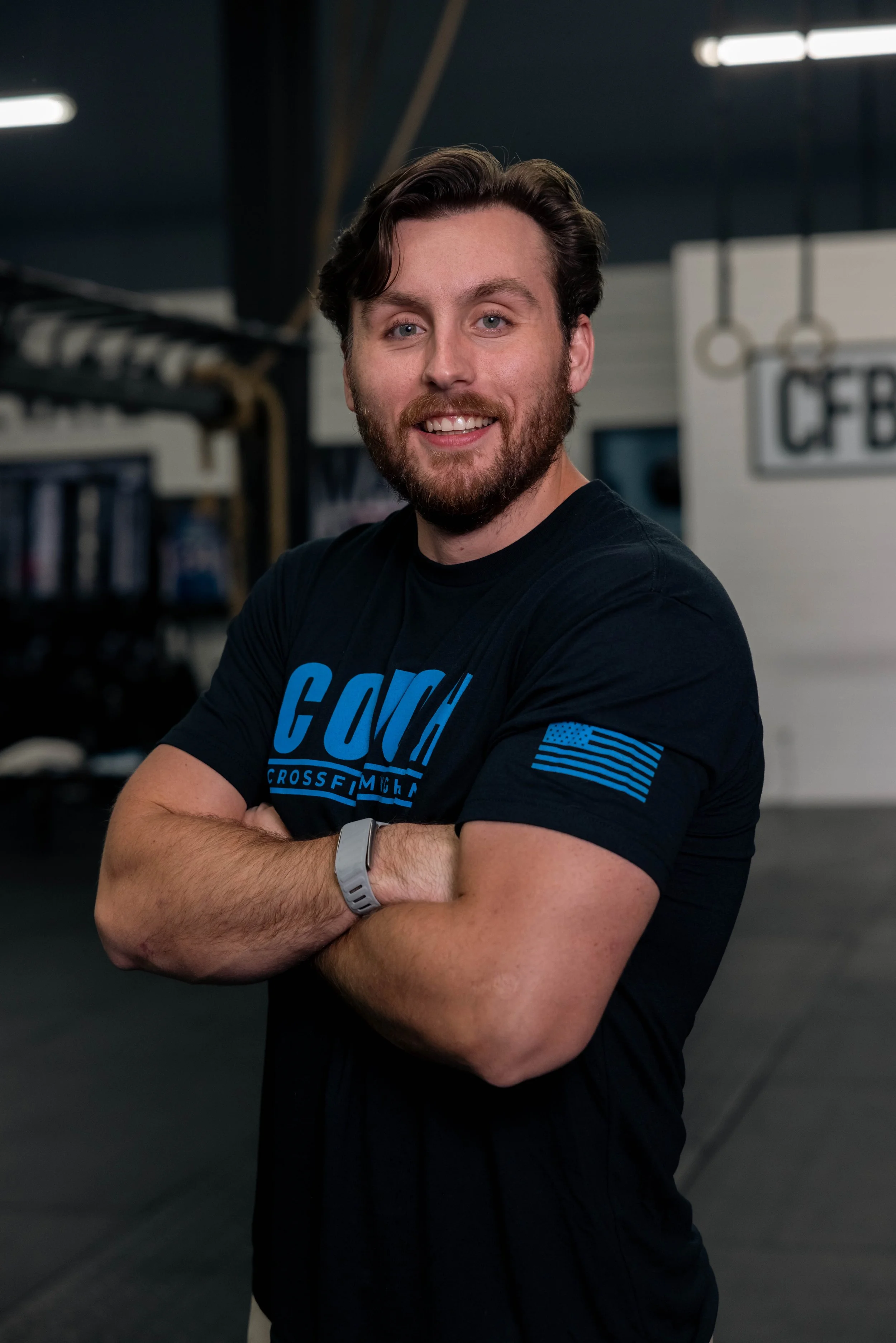 A man with a beard and brown hair standing in a gym, smiling with arms crossed, wearing a black t-shirt with blue lettering and an American flag patch on the sleeve.
