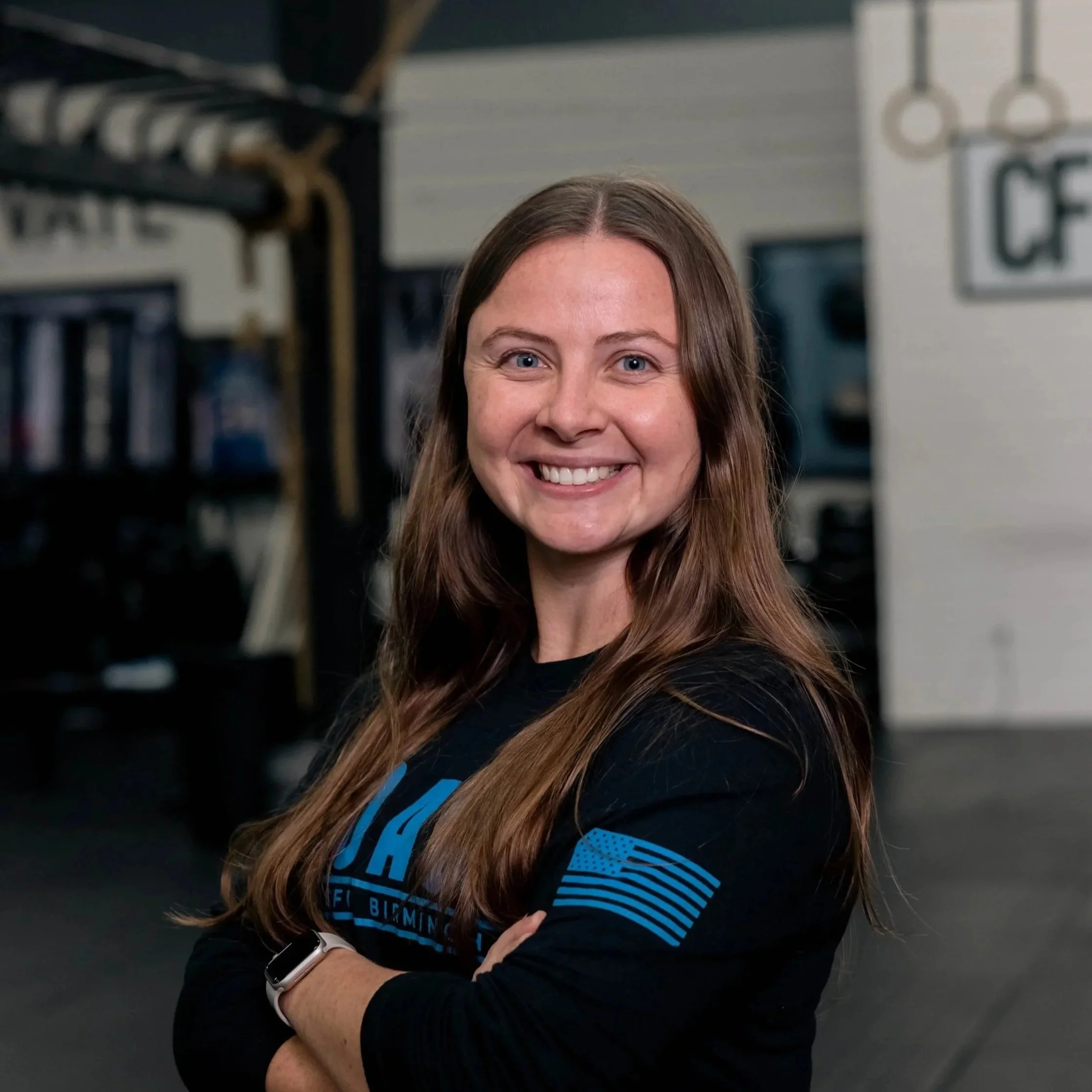 A smiling woman with long brown hair standing inside a gym, arms crossed, wearing a black shirt with blue graphics.