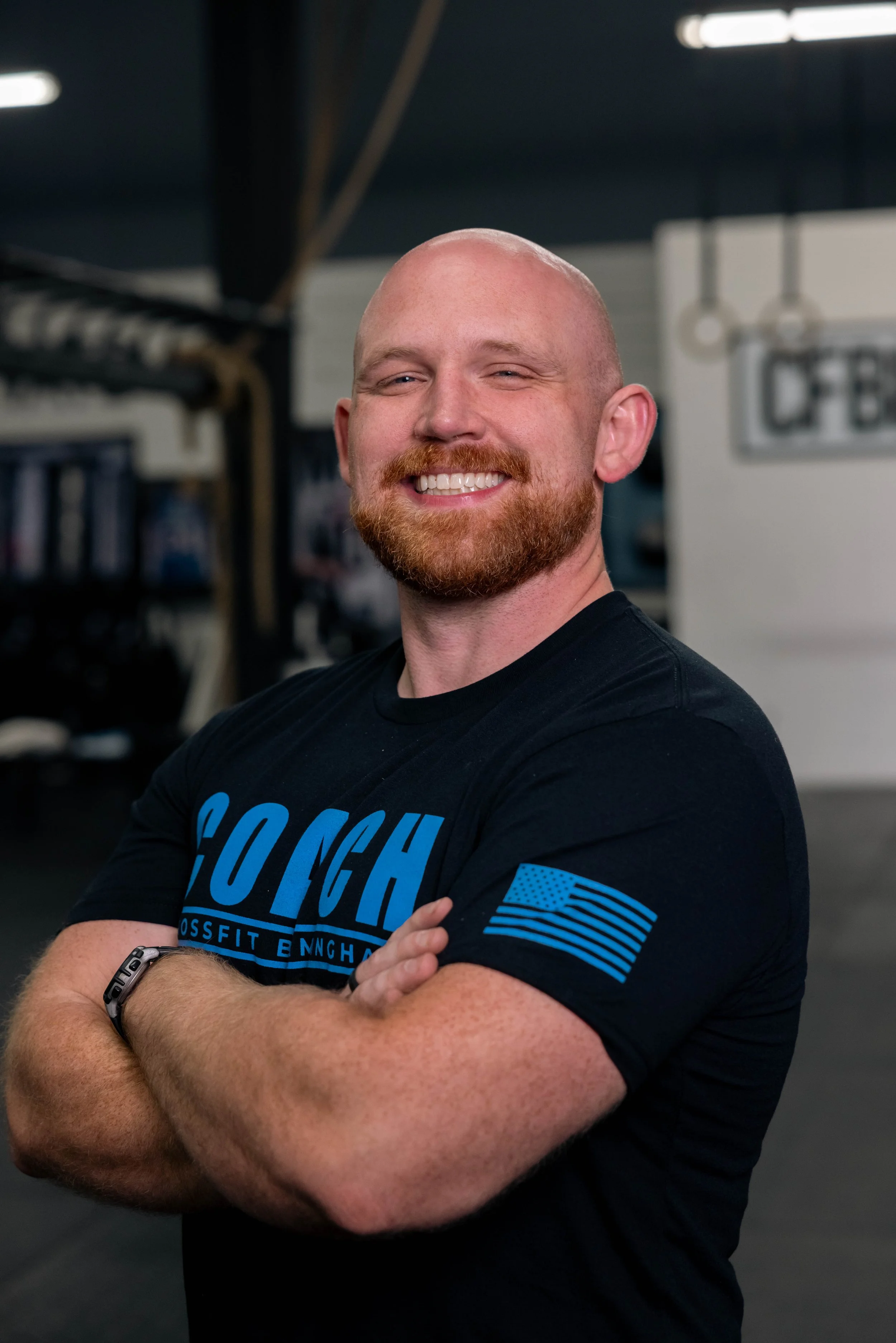 A smiling man with a beard and balding head standing with arms crossed in a gym, wearing a black t-shirt with blue lettering and an American flag patch on the sleeve.