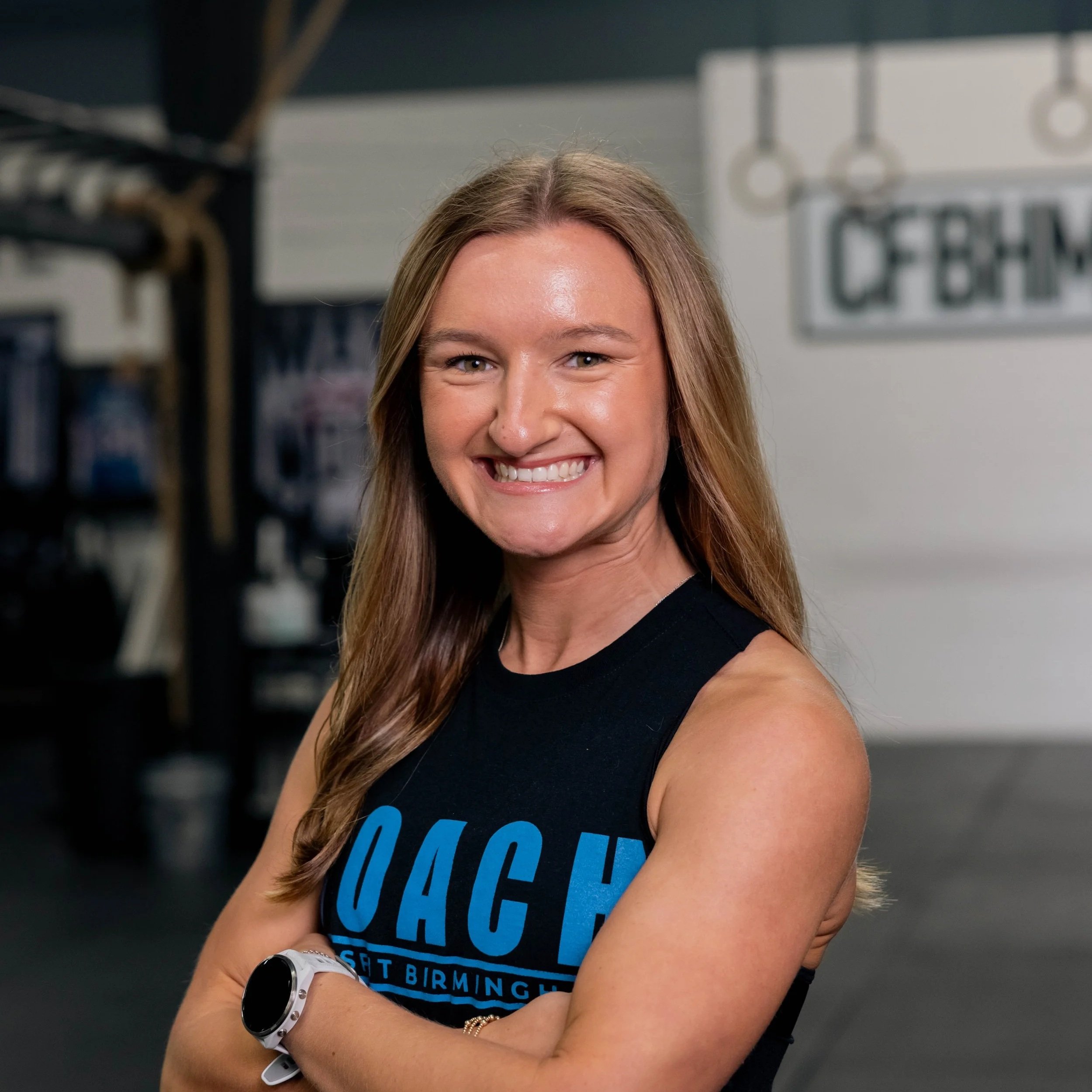 A smiling woman with long brown hair in a gym wearing a black tank top with blue text.
