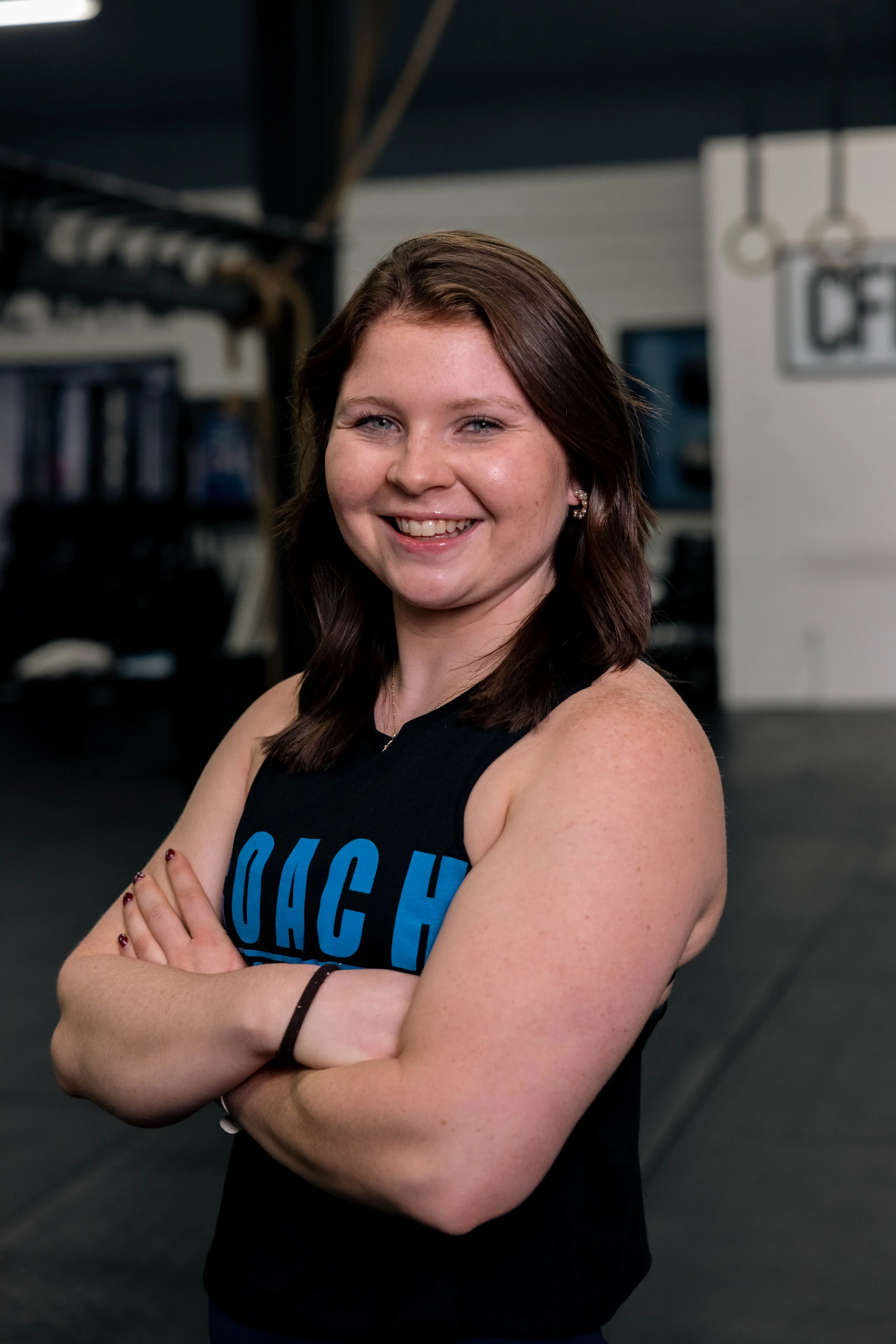 A smiling woman with brown hair and light skin, wearing a black tank top with blue letters, standing with her arms crossed inside a gym.