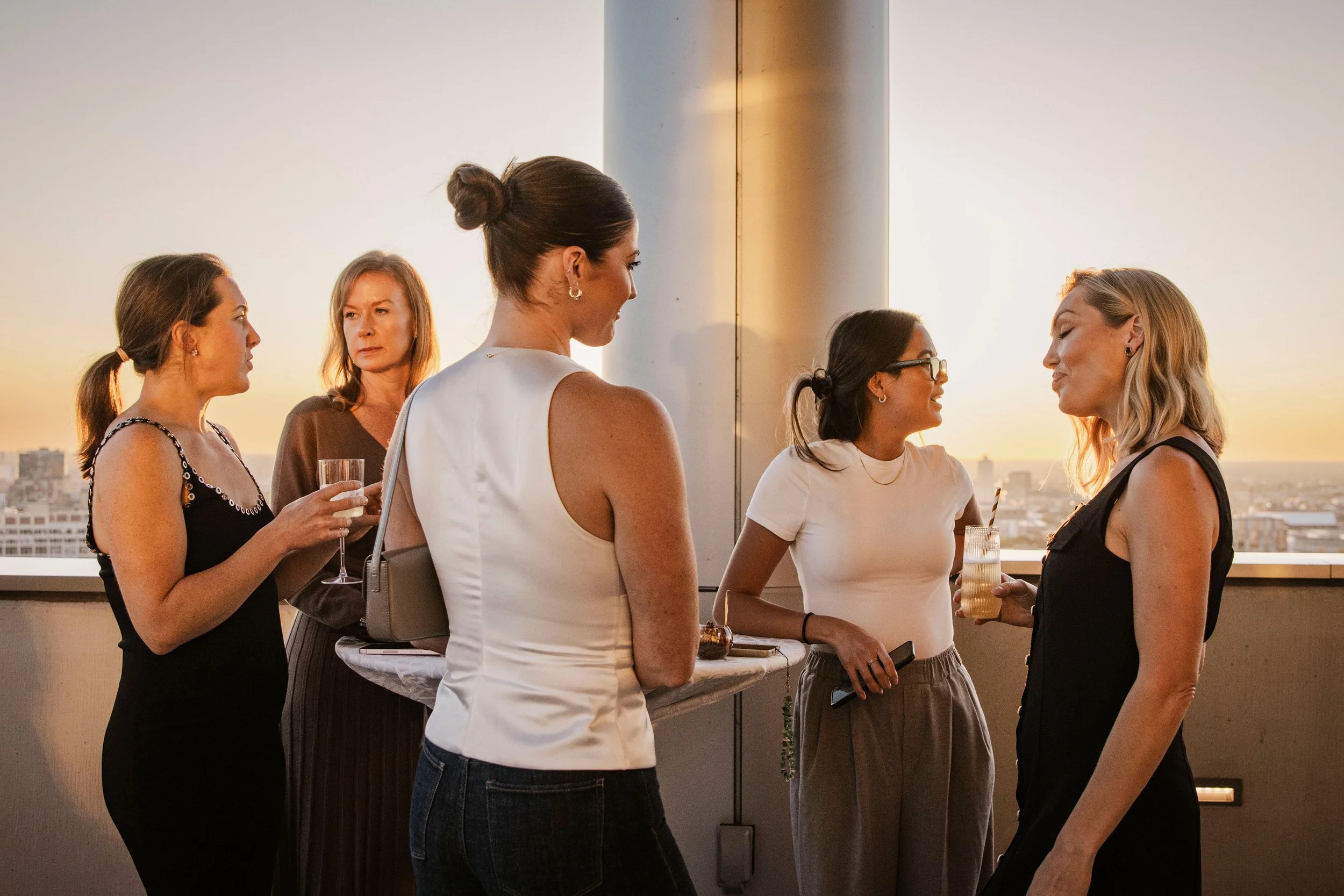 Five women at a rooftop gathering during sunset, engaged in conversation with drinks in hand.