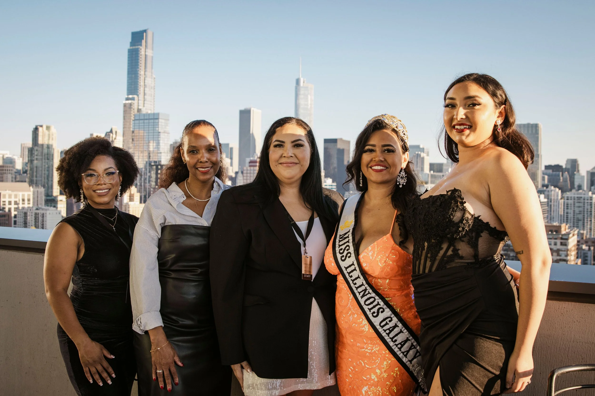 A group of five women standing on a rooftop with a city skyline in the background. One woman is wearing a sash that says 'Miss Texas Galaxy' and a crown.
