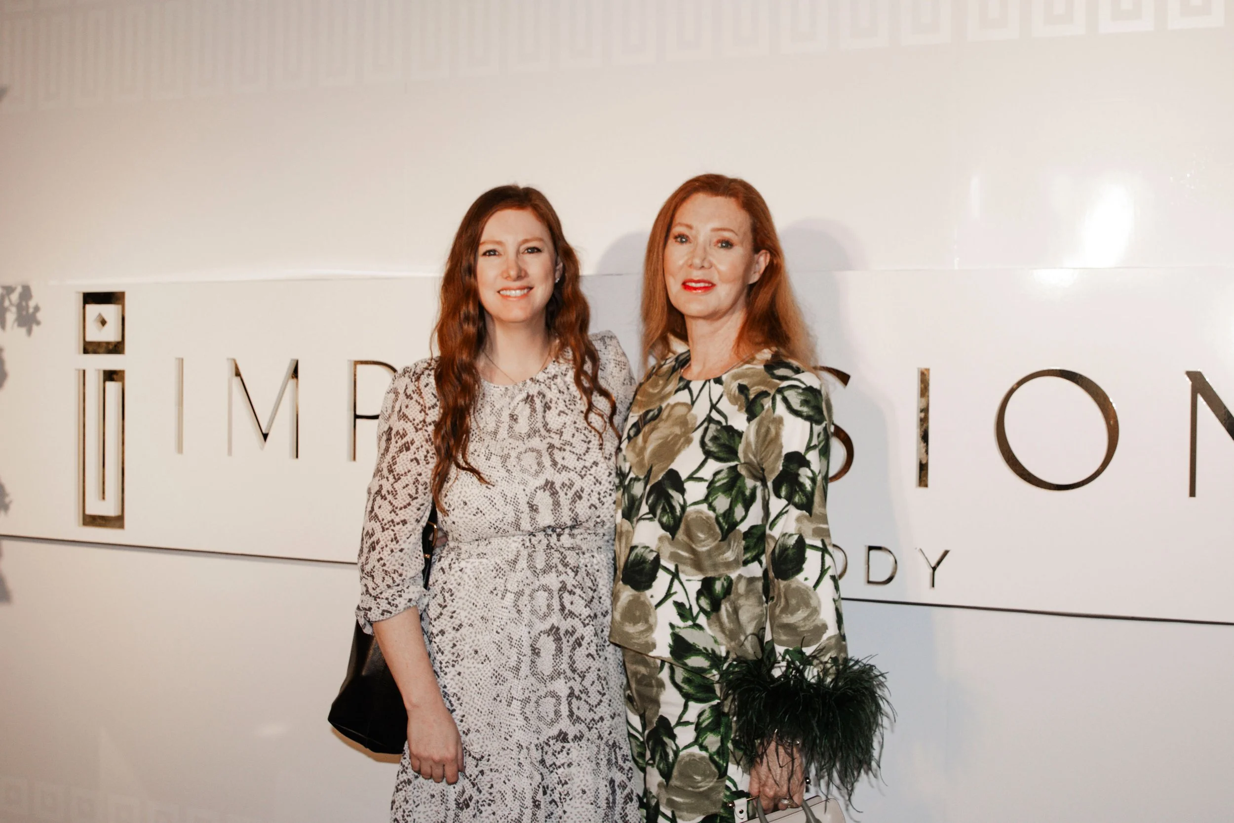 Two women standing in front of a white wall with the text 'IMPRESSION BODY', smiling for the photo, one is wearing a patterned dress, the other a floral dress. One woman is holding a purse with feathers.