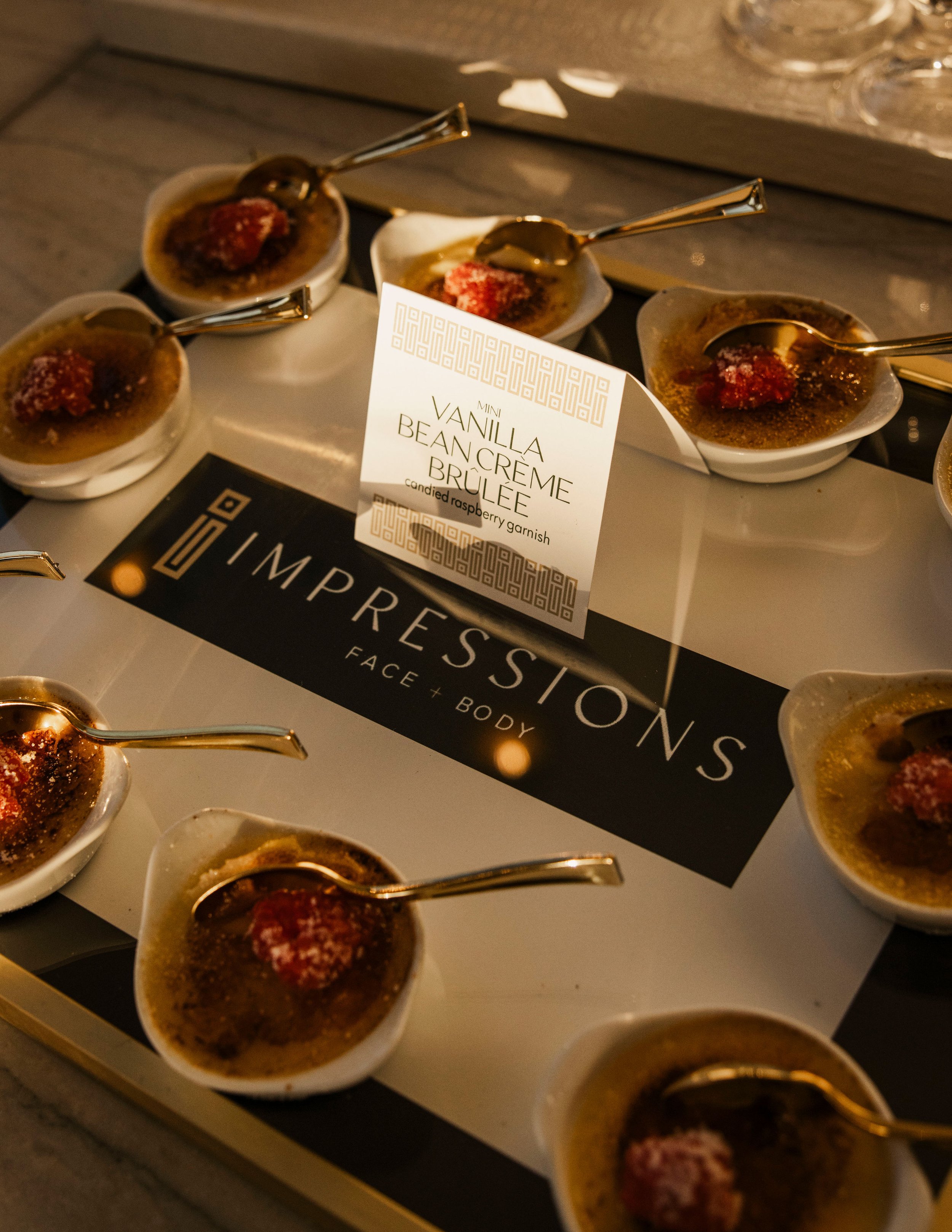 Small bowls of vanilla bean crème brûlée topped with candied raspberry garnish, arranged on a tray with a sign that reads "Mini Vanilla Bean Crème Brûlée" and a larger sign for 'Impressions Face + Body'.