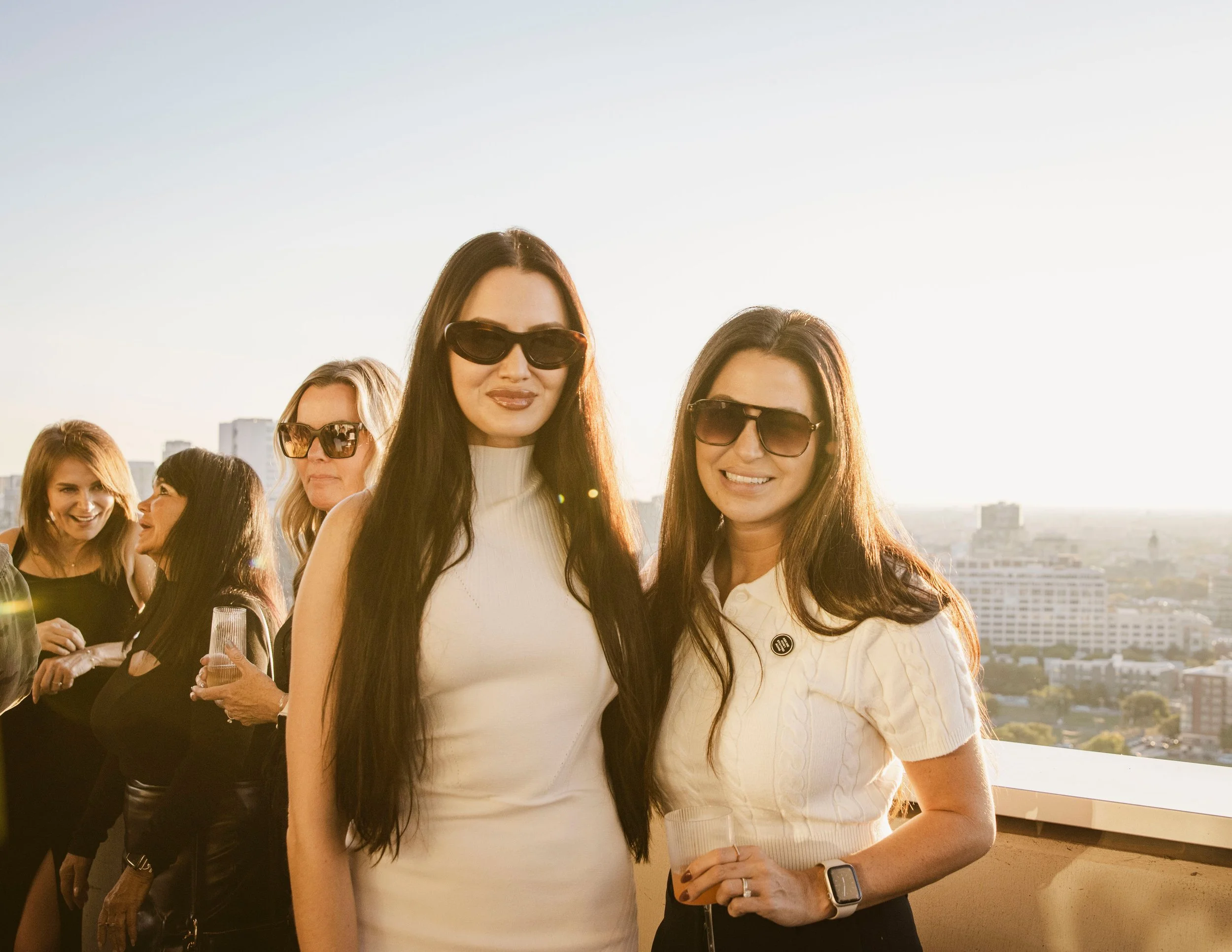 Group of women at a rooftop party during sunset, wearing sunglasses, smiling, with cityscape in background.