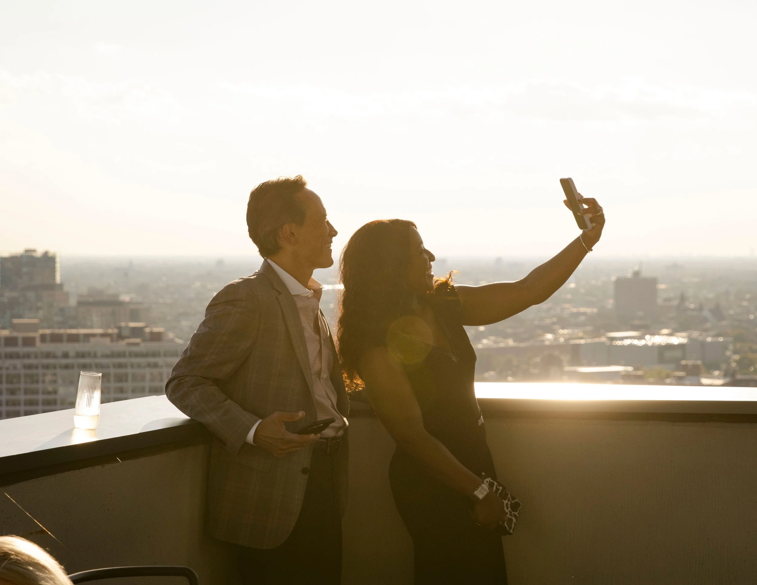 A man and woman taking a selfie on a rooftop with a cityscape in the background during sunset.
