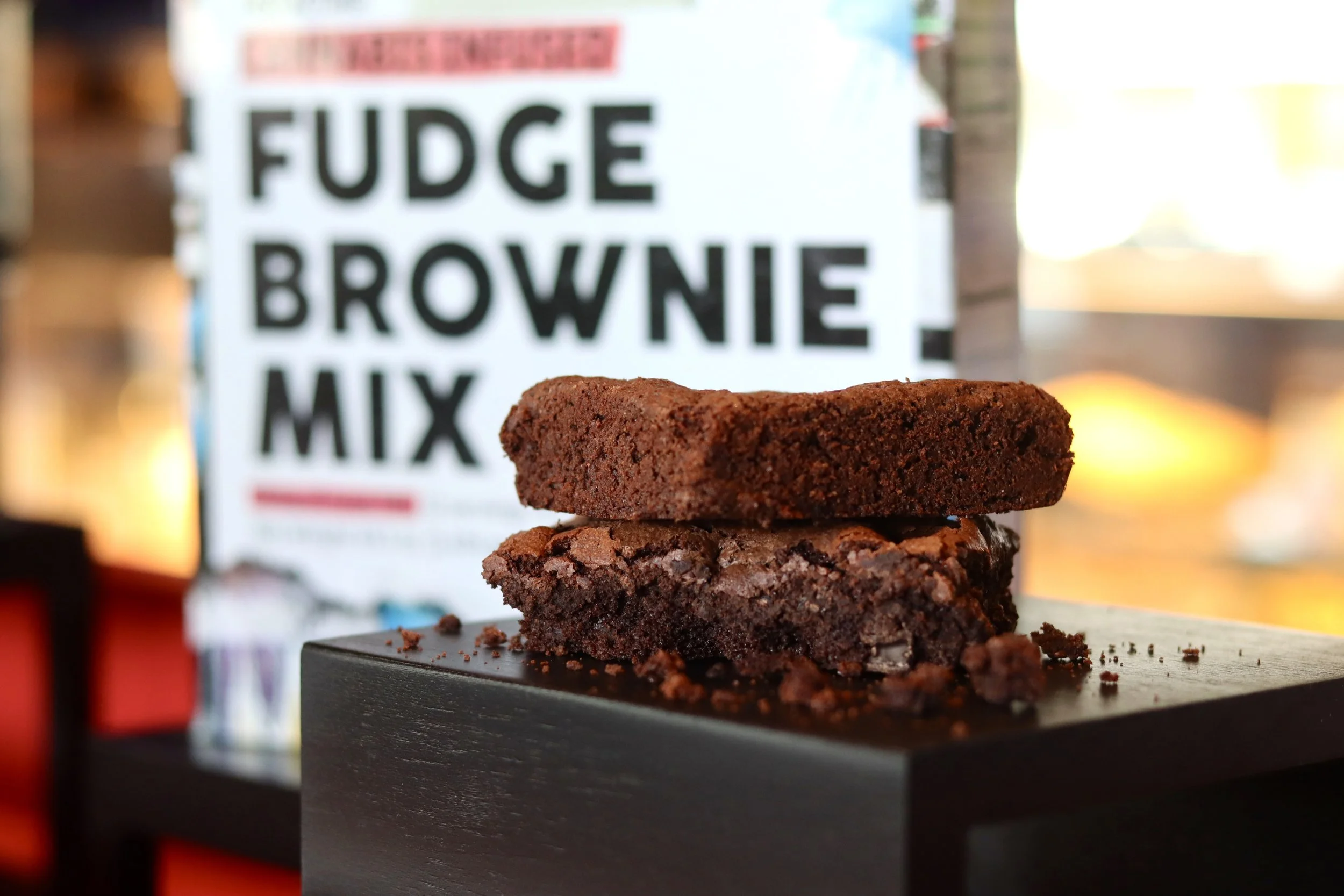 Two stacked chocolate brownies on a black display stand with a blurry baking mix package in the background.