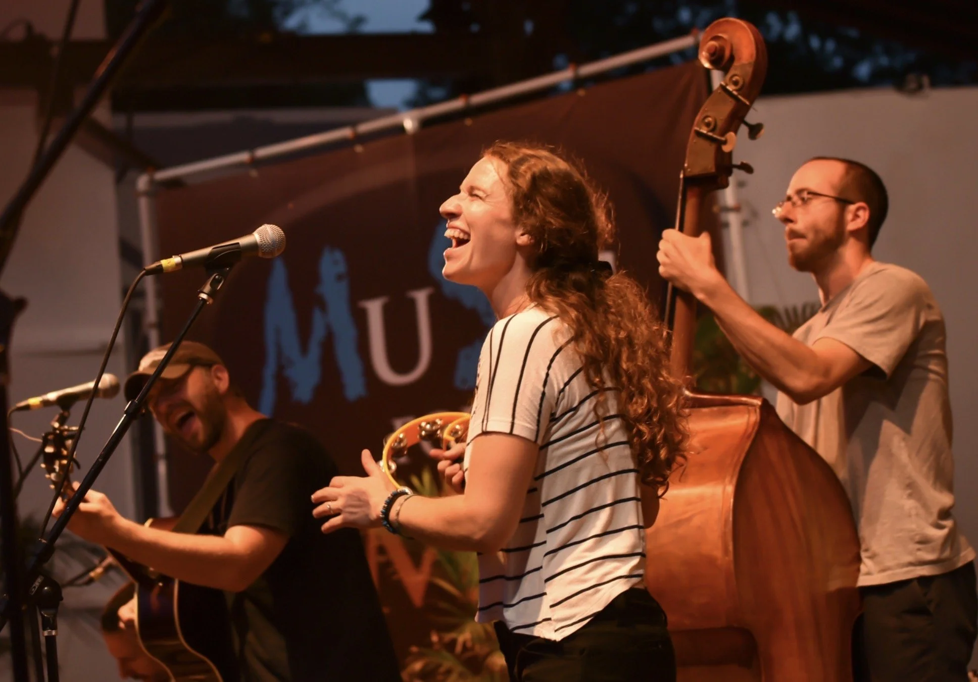 Band performing on stage with a woman singing and playing tambourine, a man playing guitar, and another playing double bass.