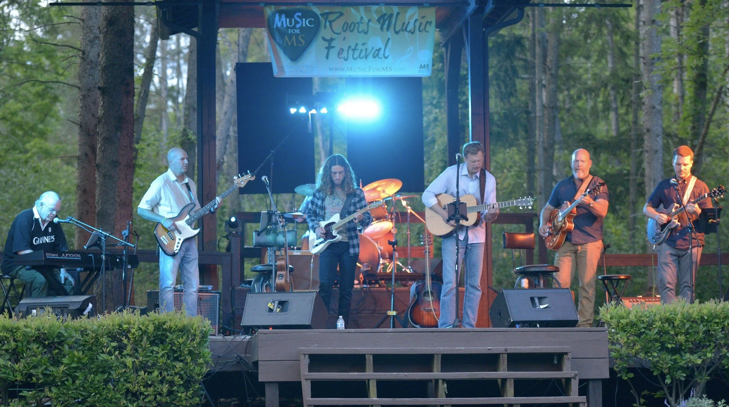 Band performing on an outdoor stage at the Music for MS Roots Music Festival, with musicians playing keyboard, bass, electric guitars, and acoustic guitar. Background of trees and lights.