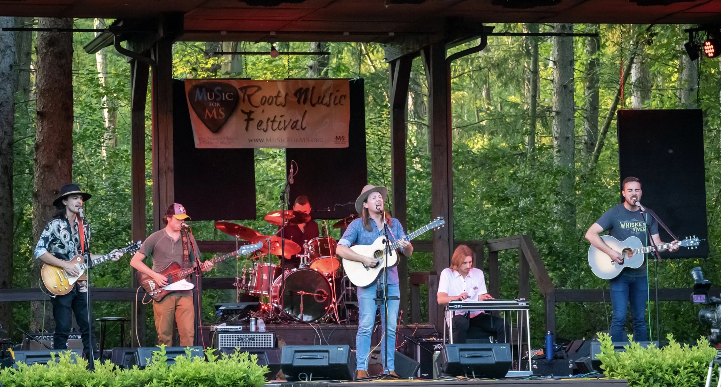 Band performing at an outdoor music festival with multiple musicians playing guitars and drums on stage, surrounded by a forest setting.