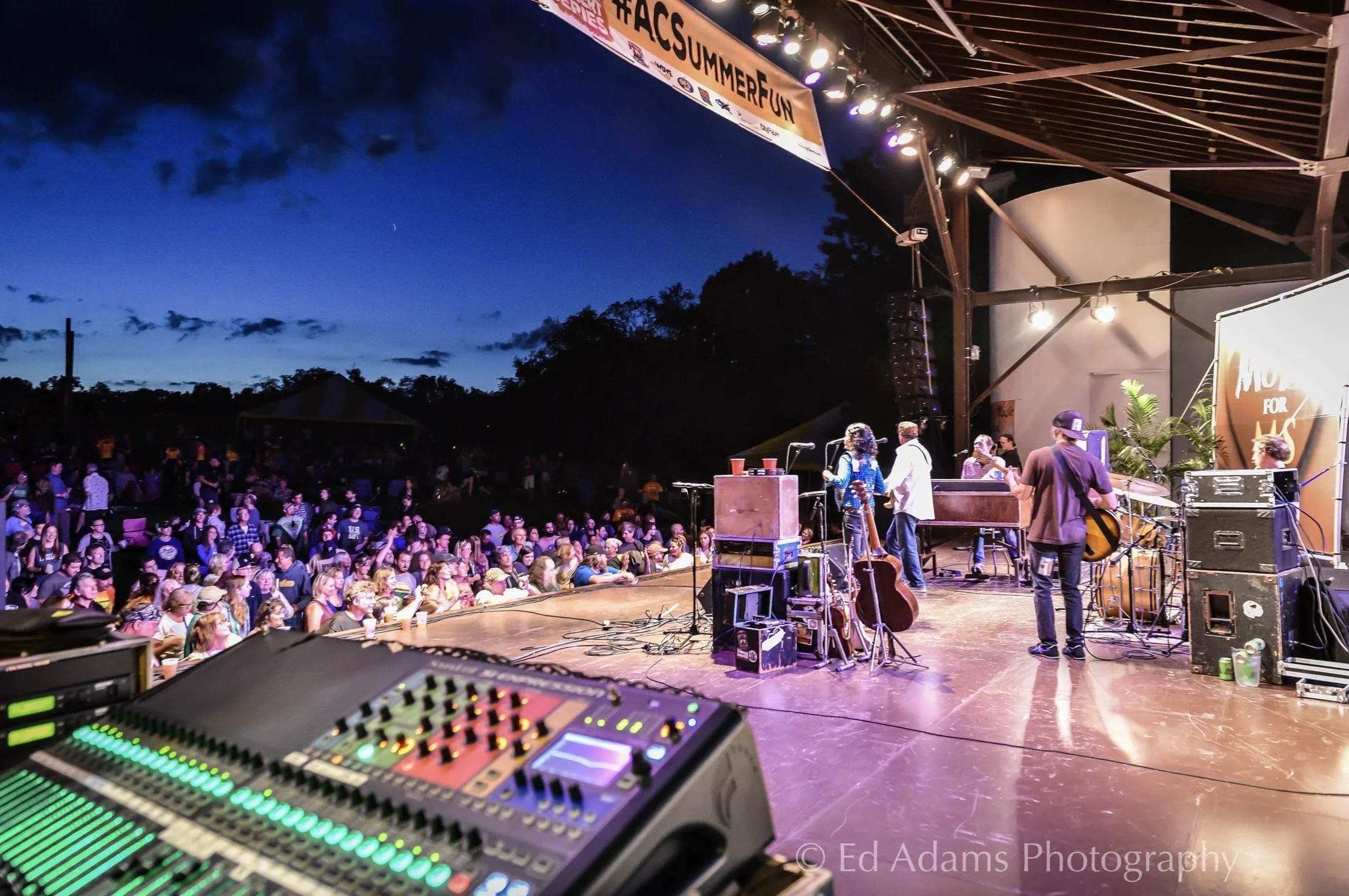 Outdoor concert stage with musicians performing to a large crowd at dusk. Sound equipment is visible in the foreground.