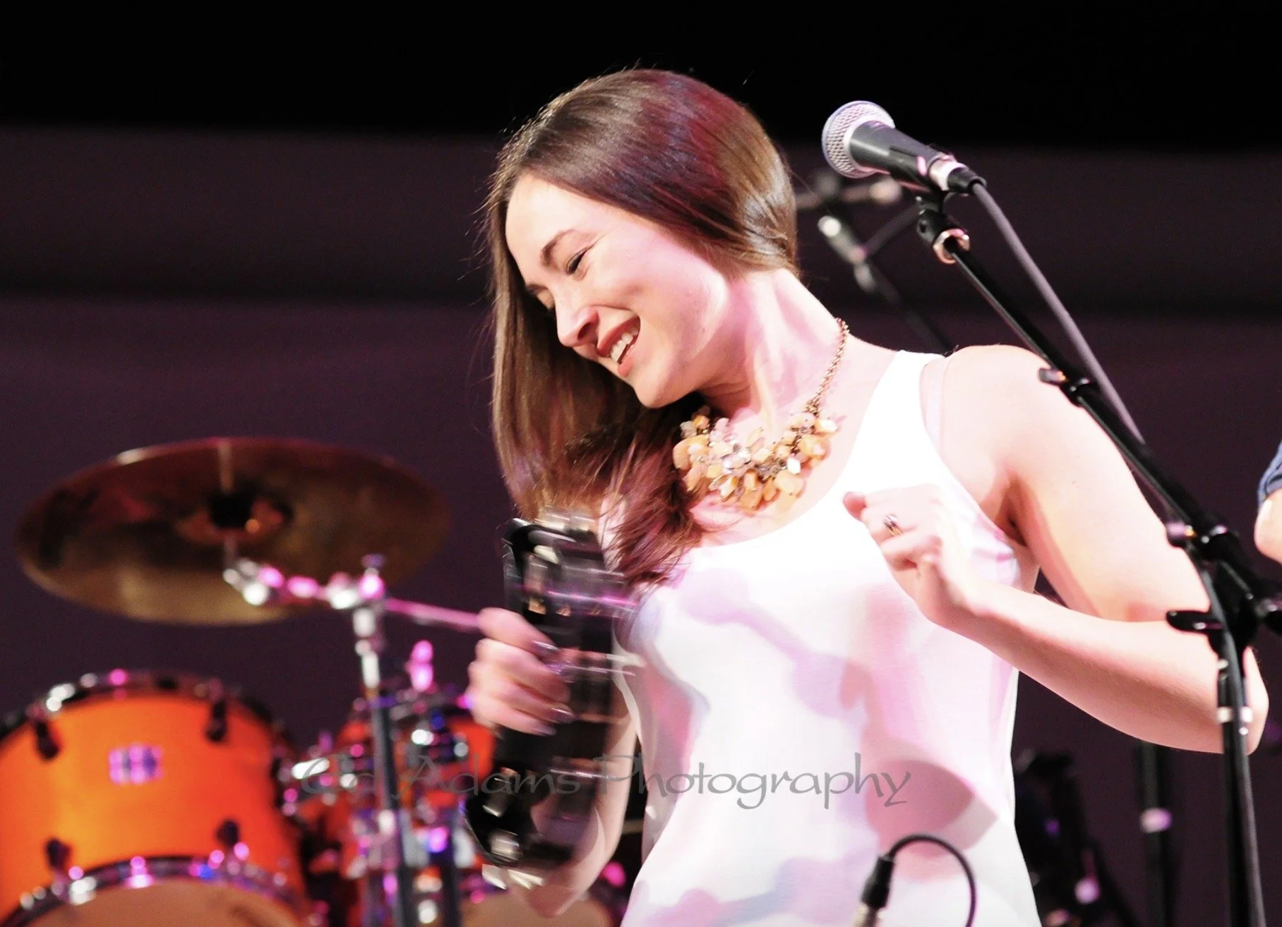 Woman in white dress smiling, holding a tambourine near a microphone on stage, with drums in the background.