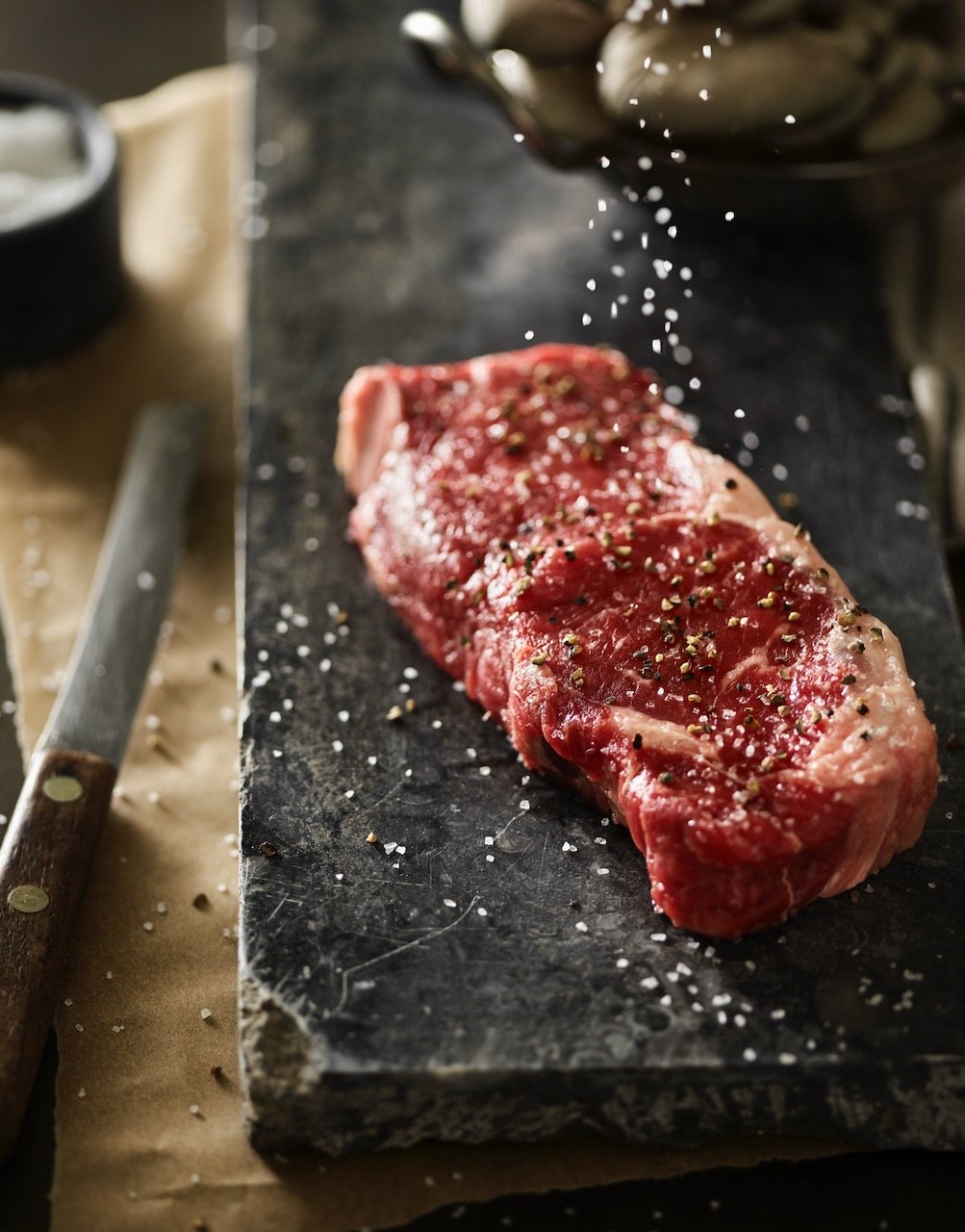 Raw steak seasoned with salt, pepper, and herbs on a black stone cutting board.