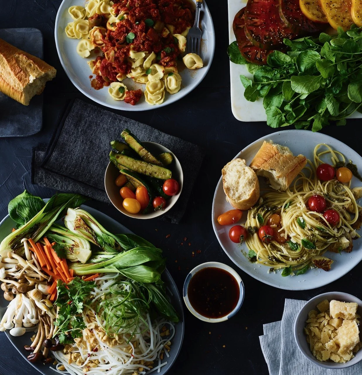 An assortment of dishes including a vegetable platter, cherry tomato salad, pasta with cherry tomatoes and basil, and a plate of tortellini with tomato sauce, alongside small bowls of soy sauce, parmesan cheese, and pickled vegetables, all arranged o