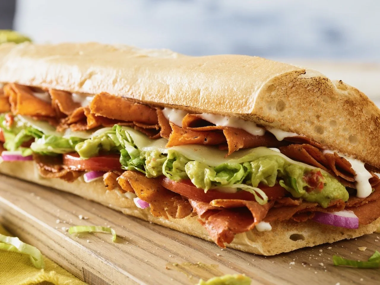 Close-up of a Quiznos sub, cured meats, lettuce, tomato, cucumber, red onion, avocado, and mayonnaise, on a wooden surface. Food styled in Denver, Colorado by food stylist Beth Hawkins of Hawkins and Company