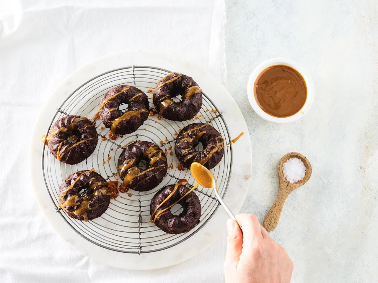Chocolate donuts drizzled with caramel on a cooling rack, with a cup of caramel sauce, a wooden spoon of salt, and a hand holding a spoon of caramel nearby.