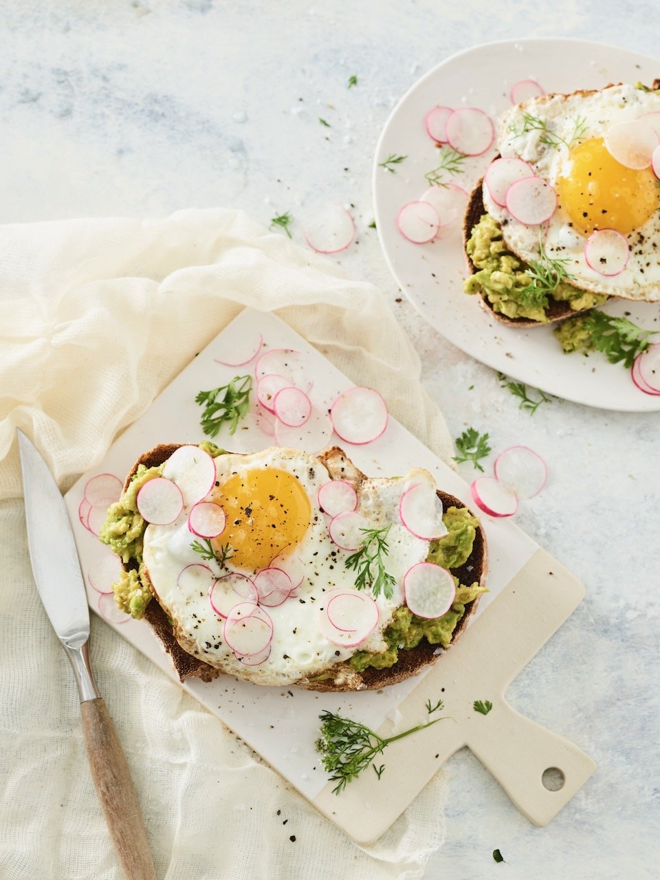 Open-faced avocado toast with a fried egg on top, garnished with radish slices and herbs, on a white cutting board and plate. Food styled in Denver, Colorado by food stylist Beth Hawkins of Hawkins and Company