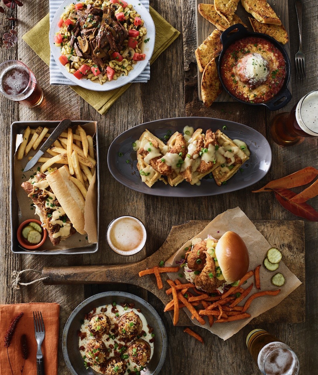 Various dishes of American comfort food on a rustic wooden table, including a cheeseburger with fries, fried chicken sandwich with sweet potato fries, pasta with chicken and cheese, loaded nachos, a meatball dish, beef stew, and a dish of beef and no