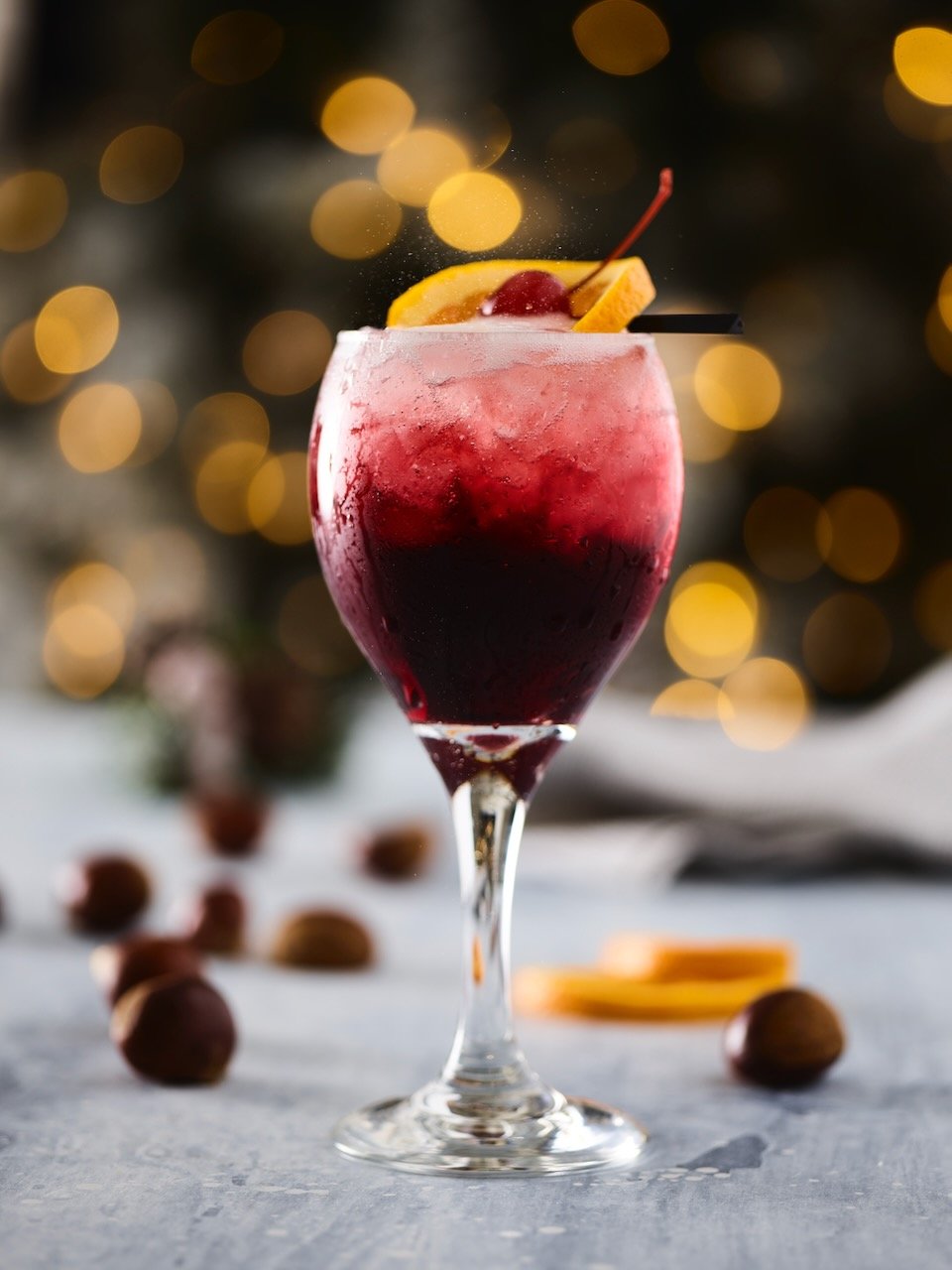 A glass of red wine cocktail garnished with an orange slice and cherry, with blurred holiday lights in the background.