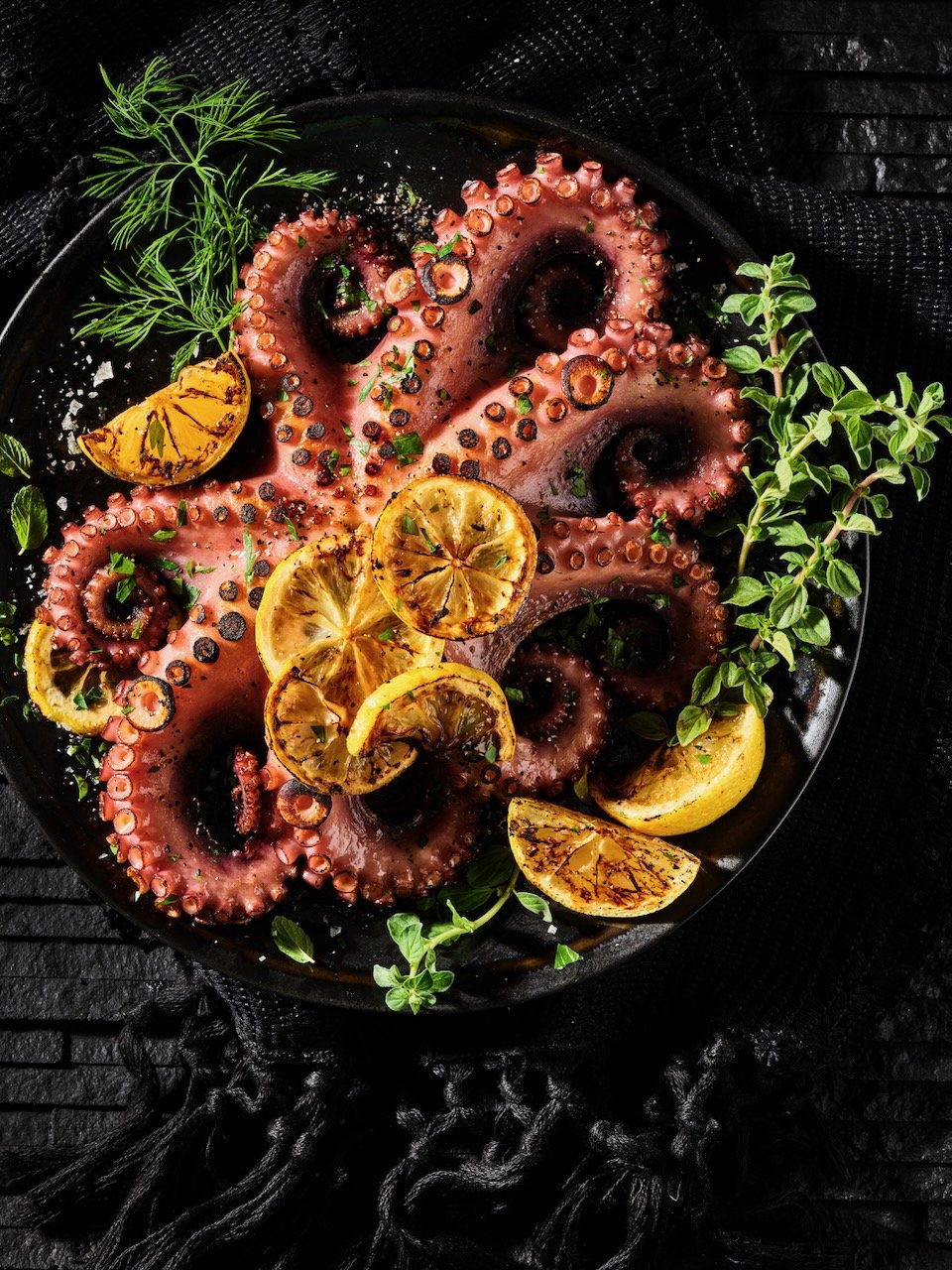 Grilled octopus tentacles on a black plate garnished with lemon slices, fresh herbs, and greens, served on a dark textured surface. Styled by Hawkins and Company's Sara Kaluzshner in Denver, Colorado Food Stylist