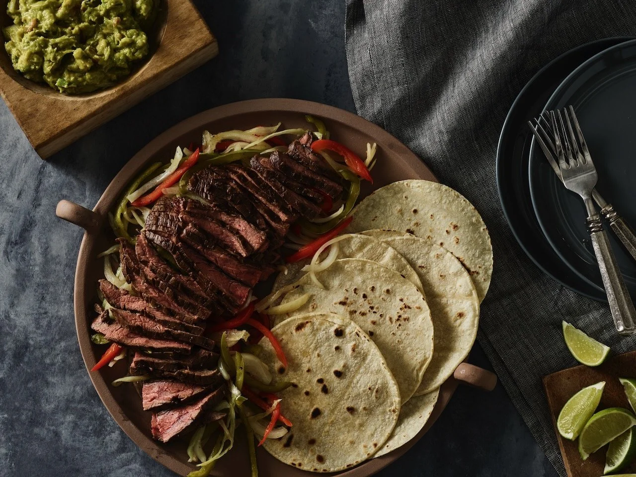 A plate with sliced steak on a bed of sautéed vegetables, served with tortillas. Guacamole is in a bowl on a wooden board in the background. Lime wedges are on a small wooden cutting board beside the plate. Silverware is placed on a black dish to the