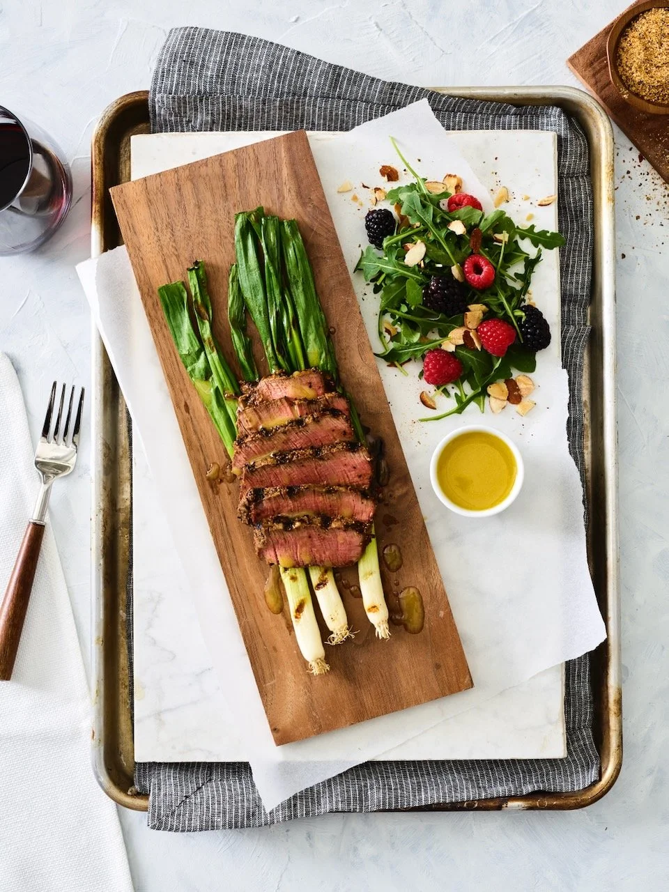 Grilled steak slices with green onions on a wooden board, side salad with mixed berries and nuts, and a small dish of olive oil on a tray