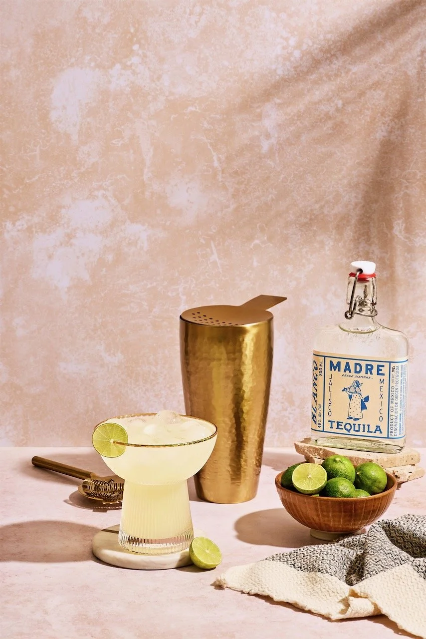 A cocktail glass with a lime wedge, a Mason jar of lime juice, a bowl of limes, a gold cocktail shaker, and a bottle of tequila on a light pink surface with a light beige textured background.