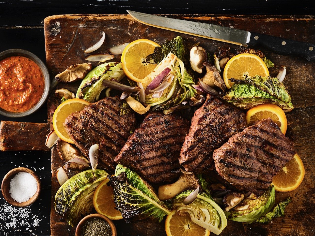 Grilled steaks with lemon slices, roasted mushrooms, and grilled lettuce on a rustic wooden table with dipping sauces. Food Stylist Colorado Food Stylist Denver. Hawkins and Company Beef It's What's for Dinner is the client.