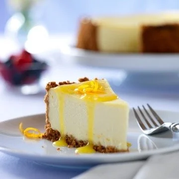 A slice of lemon cheesecake on a white plate with a fork, with a blurred background of a whole cheesecake and berries.