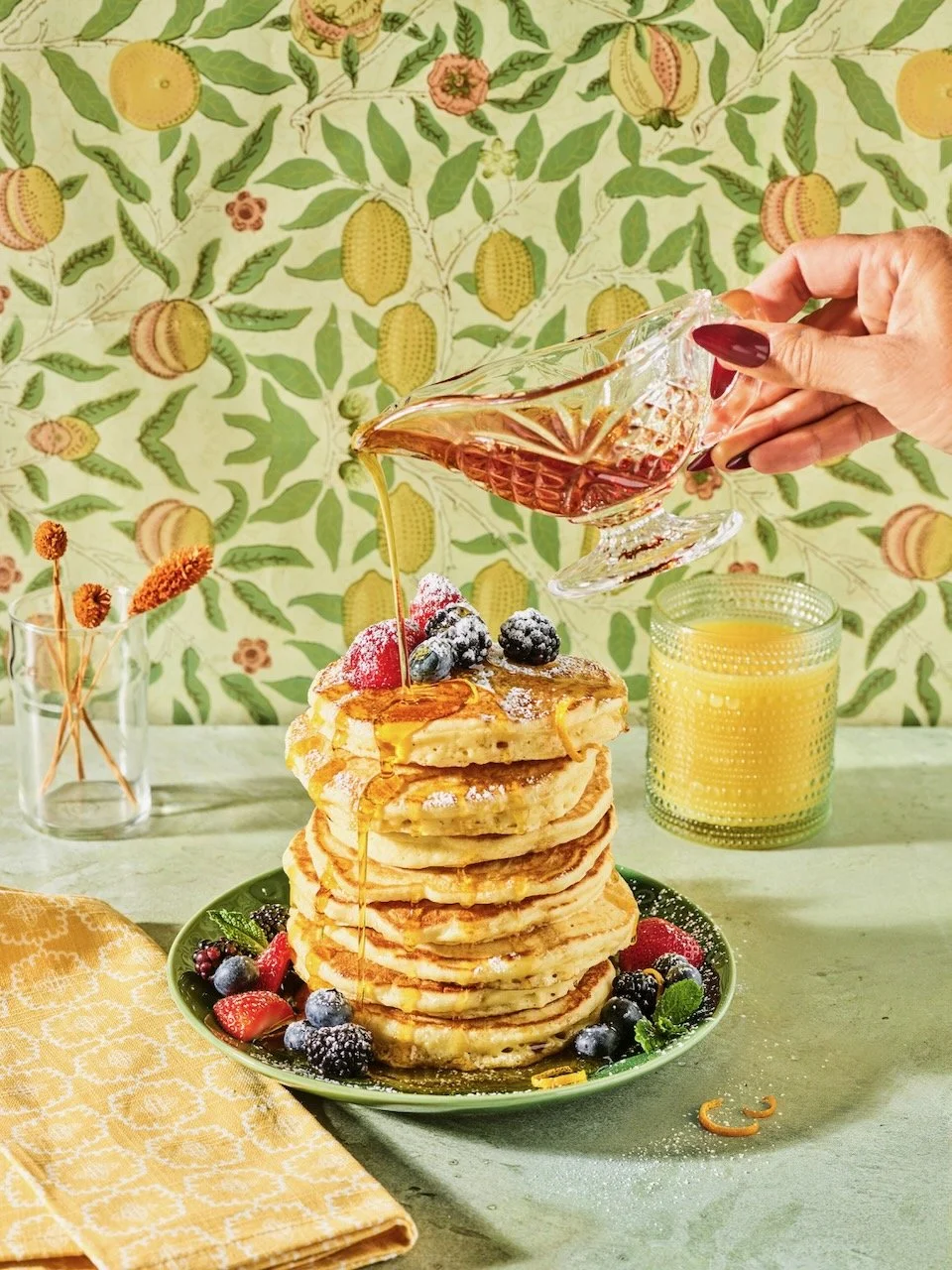 Stack of seven pancakes topped with fresh berries and powdered sugar, with syrup being poured from a glass pitcher, on a green plate with additional berries around it. In the background, there are a vase with dried flowers, a yellow drink in a textur