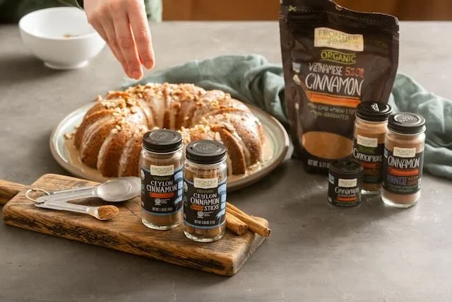 A bundt cake on a white plate with cinnamon sticks and a small spoon on a wooden cutting board. Several jars of Ceylon cinnamon and a packet of Vietnamese cinnamon are nearby on a gray surface.
